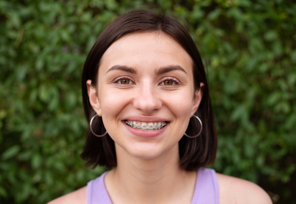 A smiling woman with hoop earrings and braces in front of a tree