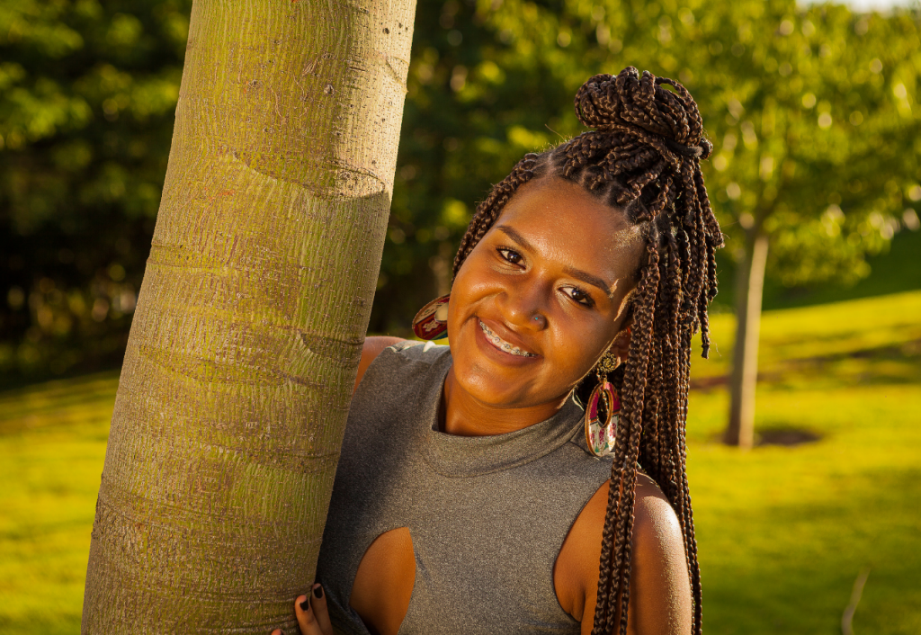 A young woman with braces peeks out from behind a palm tree trunk