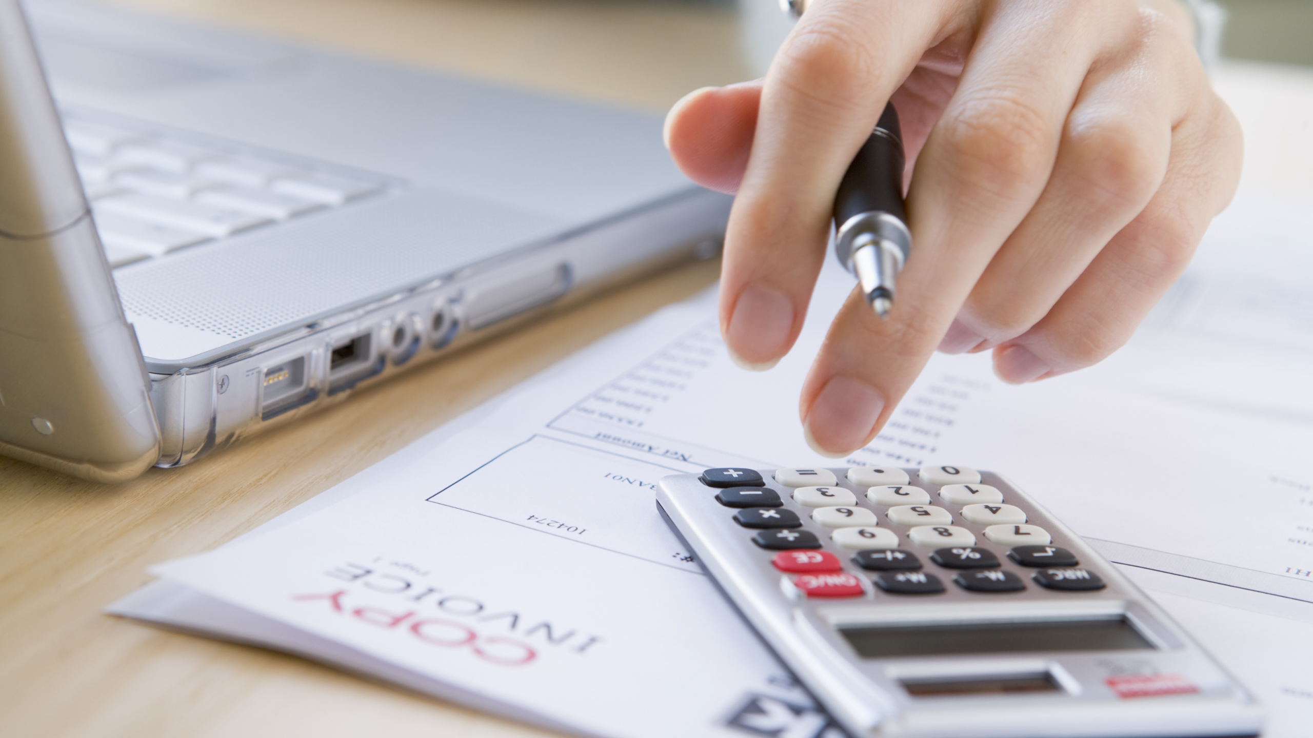 1 A close-up shot of a hand with a pen, hovered over a calculator on top of a pile of paperwork next to an open laptop.