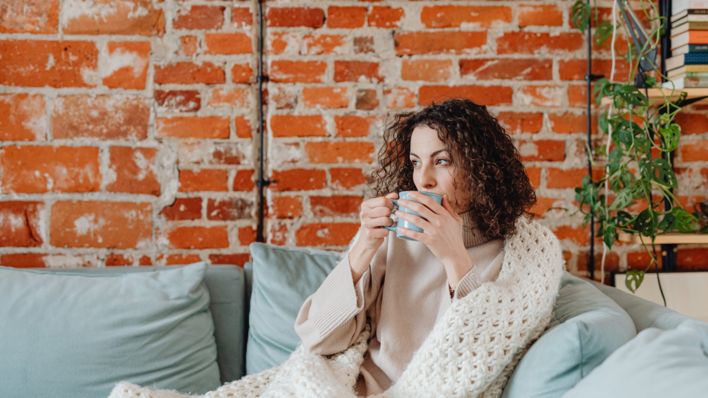 A curly-haired woman sits on the couch sipping a hot beverage