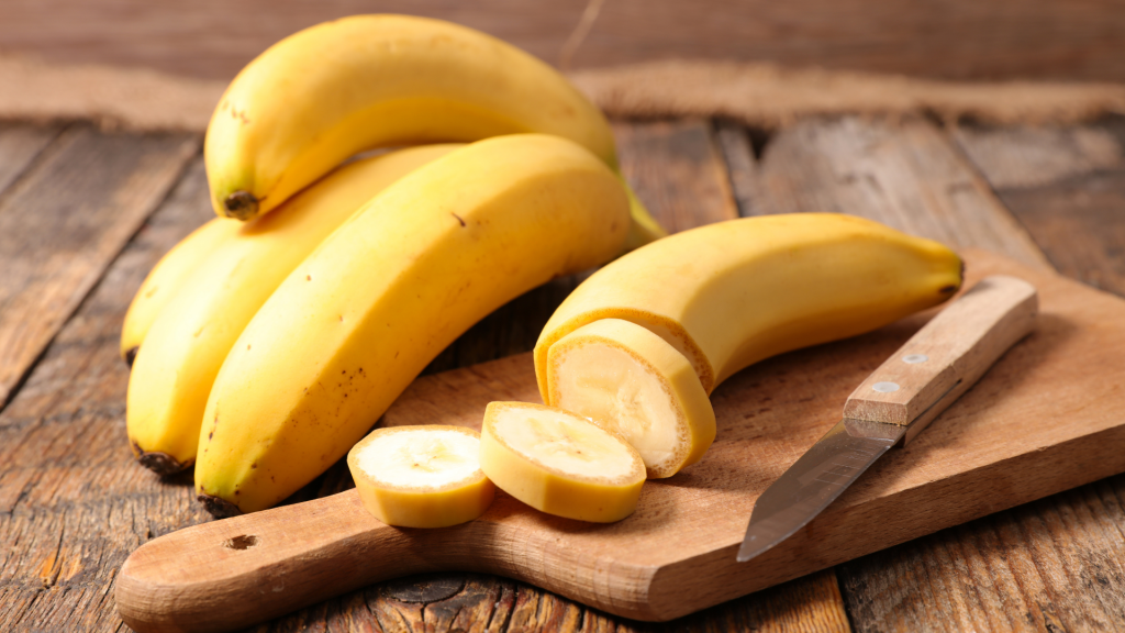 Several bananas sit next to a knife on a cutting board, one has been sliced several times.